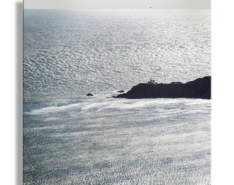 May include: A black and white photograph of a rocky coastline with a lighthouse in the distance. The ocean is calm and the water is reflecting the sunlight.