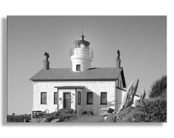 May include: A black and white photograph of a lighthouse with a white tower and a white building. The lighthouse is on a hill with trees and bushes in the background.