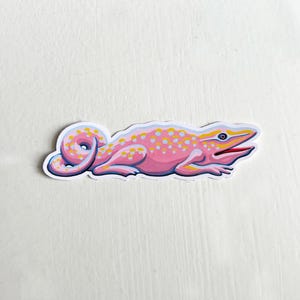 Pode incluir: Um adesivo de lagarto rosa e amarelo com manchas brancas. O lagarto está enrolado com a cauda na boca.