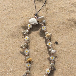 May include: A white and gold phone charm with a seashell and daisy design. The charm is on a sandy background.