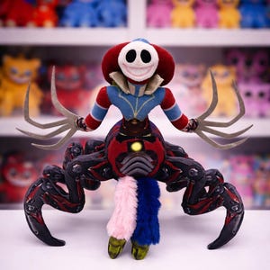 Puede incluir: Un peluche con una cara sonriente, ropa roja y azul y múltiples brazos. El juguete tiene una cabeza roja con una cara blanca y una amplia sonrisa cosida. El cuerpo es rojo y negro con una cola peluda rosa, azul y blanca. Otros peluches están en el fondo.