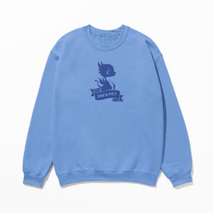 May include: Light blue sweatshirt with a dark blue graphic of a cartoon creature holding a banner that reads "DM'S PET". The sweatshirt has a crew neck and long sleeves. The graphic is centered on the chest.