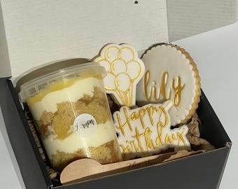 May include: A black gift box with a clear container of lemon cake, a wooden spoon, and three white cookies with gold lettering. The cookies say "Happy Birthday", "Lily", and "Lemon".