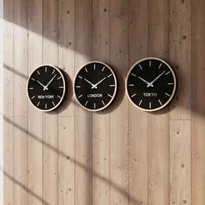 Peut inclure: Trois horloges murales noires avec des aiguilles blanches et des fuseaux horaires : New York, Londres et Tokyo. Chaque horloge a un cadre doré et est fixée sur un mur en bois.
