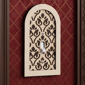 Könnte beinhalten: Eine dekorative Lichtschalterabdeckung mit gewölbtem Oberteil und cremefarbenem Rahmen. Die Abdeckung zeigt ein lasergeschnittenes Blumendesign in Dunkelbraun, das einen Standardschalter umgibt. Die Abdeckung ist an einer roten Wand angebracht.
