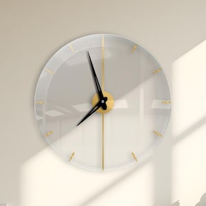 May include: A modern, clear acrylic wall clock with gold accents. The clock features gold hour markers and hands, with a black minute and hour hand. The clock is mounted on a light-colored wall, with a wooden cabinet and plant in the background.