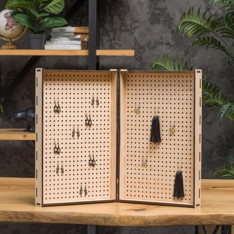 May include: A wooden earring display stand with two hinged panels. Each panel has a grid of holes for hanging earrings. The stand is open and has several pairs of earrings hanging from the holes.