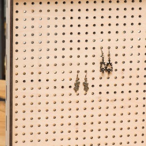 May include: A wooden pegboard with a grid of holes, used for displaying earrings. Two pairs of earrings are displayed on the board. One pair features a delicate design with a small, ornate pendant. The other pair features a simple design with a small, round pendant.