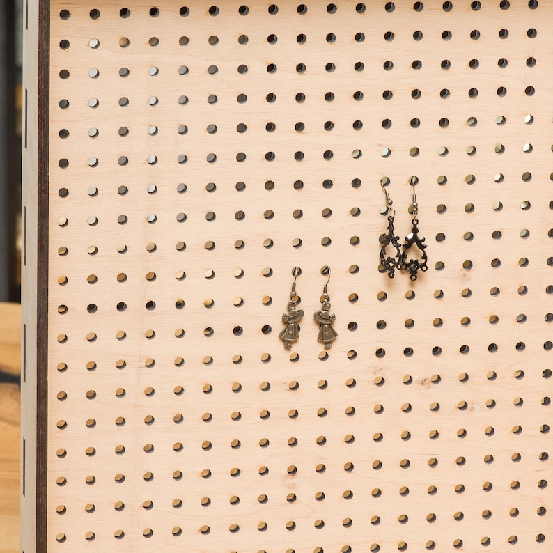 May include: A wooden pegboard with a grid of holes, used for displaying earrings. Two pairs of earrings are displayed on the board. One pair features a delicate design with a small, ornate pendant. The other pair features a simple design with a small, round pendant.