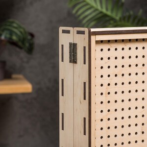 May include: A wooden cabinet with a perforated front panel and a hinged door. The cabinet is made of light wood with dark brown accents. The door has a metal hinge.