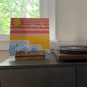 May include: A wooden record stand with the words "Now Spinning" on it. A vinyl record with a sunset and ocean scene is displayed on the stand. The record cover has the word "Corabelle" written on it.