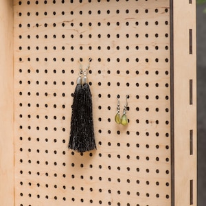 May include: A wooden earring holder with a pegboard design. Two pairs of earrings are displayed on the board. One pair is black with tassels and the other pair is green.