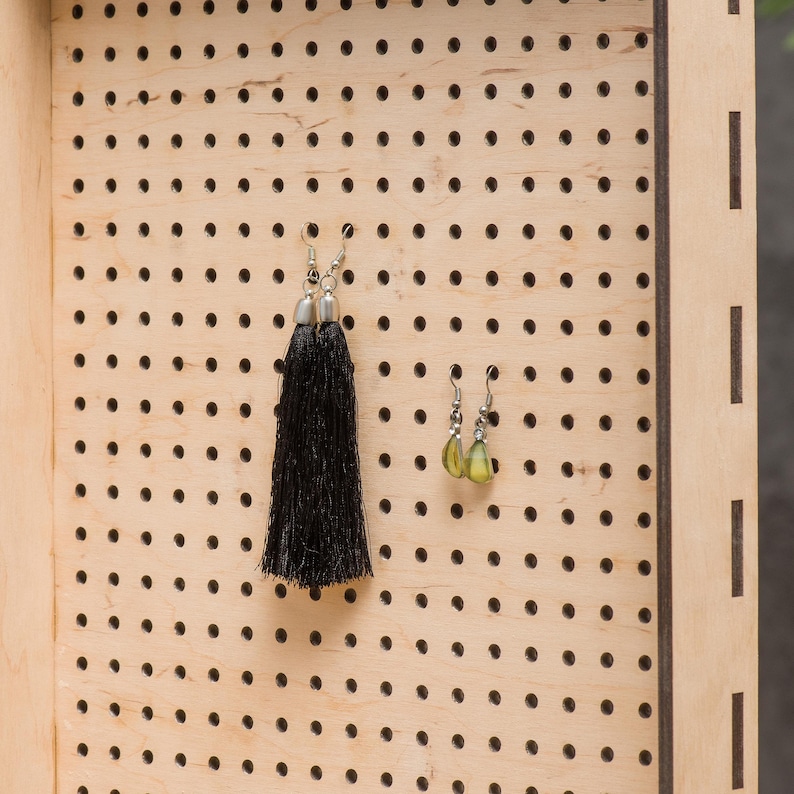 May include: A wooden earring holder with a pegboard design. Two pairs of earrings are displayed on the board. One pair is black with tassels and the other pair is green.