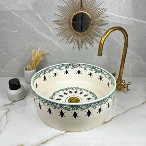 Puede incluir: Un lavabo redondo de cerámica color crema con un diseño decorativo turquesa y azul marino en el borde y el centro. Un grifo dorado y un pequeño espejo decorativo se encuentran en el fondo.