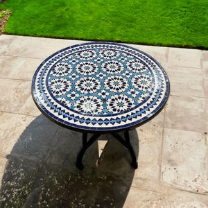 Könnte beinhalten: Ein runder, weißer Tisch mit einer blauen und weißen Mosaik-Tischplatte mit geometrischem Muster. Der Tisch hat ein schwarzes Metallgestell und steht auf einer hellgrauen, gefliesten Terrasse.