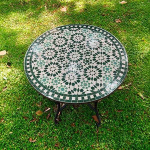 Op de afbeelding: Een ronde, groen-witte mozaïektafel met een geometrisch patroon. De tafel staat op een groen gazon.