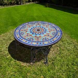 Pode incluir: Uma mesa redonda de mosaico com um padrão geométrico azul, marrom e dourado. A mesa tem uma base de metal preto e está sentada em um gramado verde.