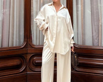 May include: Cream-colored satin two-piece set, featuring a long-sleeved button-down shirt and wide-leg pants. The outfit is worn with sunglasses and a necklace. A brown handbag is held in the hand. The background includes a dark wooden door and a patterned floor.