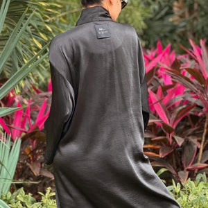 May include: A long, black satin robe with a collar and long sleeves. The robe has a small tag on the back with the text "SO by Nina". The background features lush green and pink foliage.