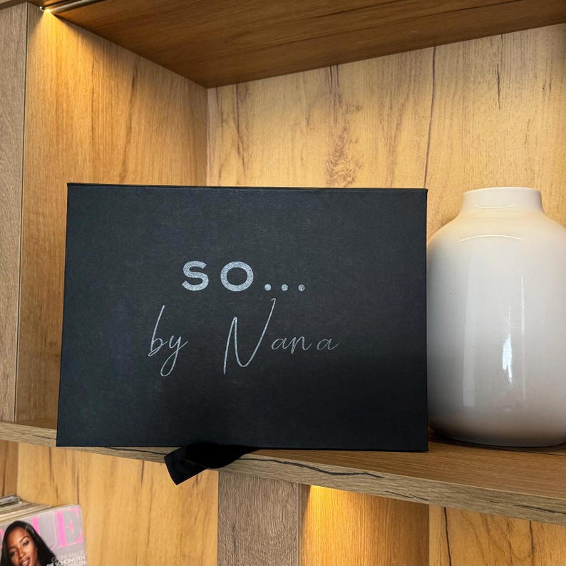 May include: A black box with the text "SO... by Nana" in silver lettering sits on a wooden shelf. A white vase is to the right of the box. The shelf is part of a wooden cabinet.