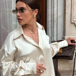 May include: A woman wearing a cream-colored satin button-down shirt, sunglasses, and pearl jewelry. She is holding a brown handbag with a gold clasp. The background features a marble wall and a window.