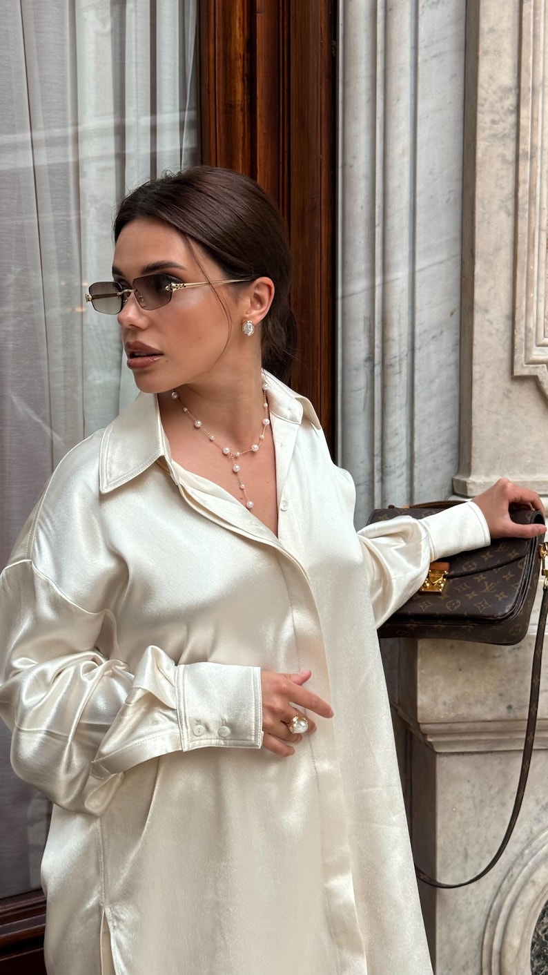 May include: A woman wearing a cream-colored satin button-down shirt, sunglasses, and pearl jewelry. She is holding a brown handbag with a gold clasp. The background features a marble wall and a window.