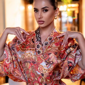 May include: A vibrant, patterned kimono-style robe with a wrap closure. The robe features a paisley design in shades of red, orange, and gold. The sleeves are wide and flowing. The robe is tied at the waist with a matching belt.