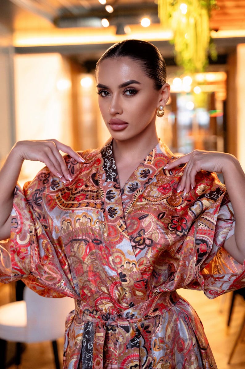 May include: A vibrant, patterned kimono-style robe with a wrap closure. The robe features a paisley design in shades of red, orange, and gold. The sleeves are wide and flowing. The robe is tied at the waist with a matching belt.