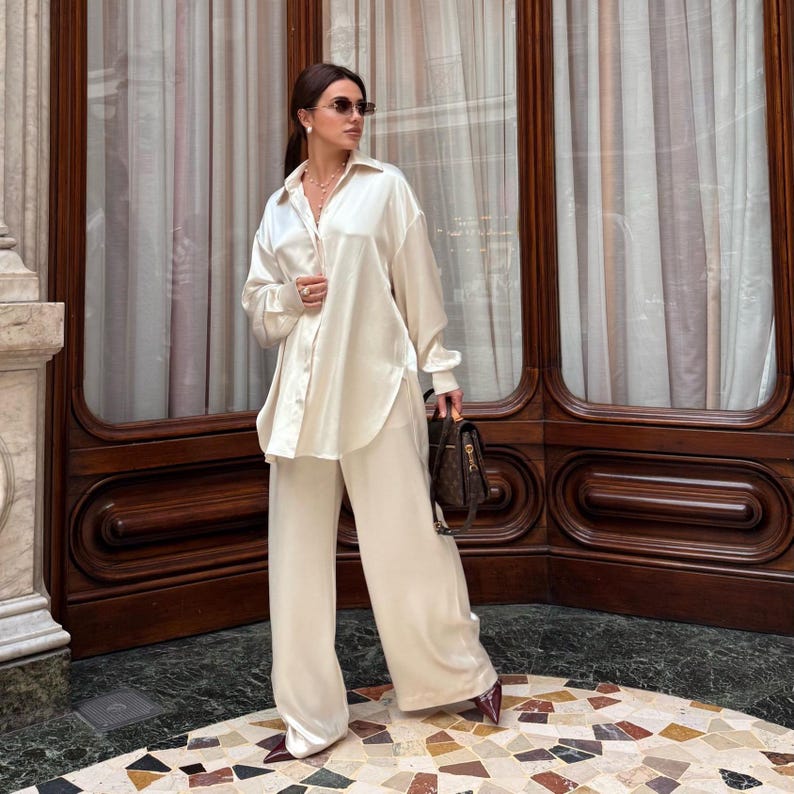 May include: Cream-colored silk shirt and wide-leg pants set. The long-sleeved button-down shirt is paired with matching trousers. The outfit is accessorized with sunglasses and a handbag. The ensemble is a stylish, monochromatic look.