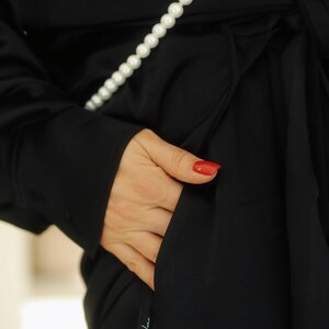 May include: A black long-sleeved shirt with a pearl necklace and a pocket on the side. The shirt has a label that says "by Uma".
