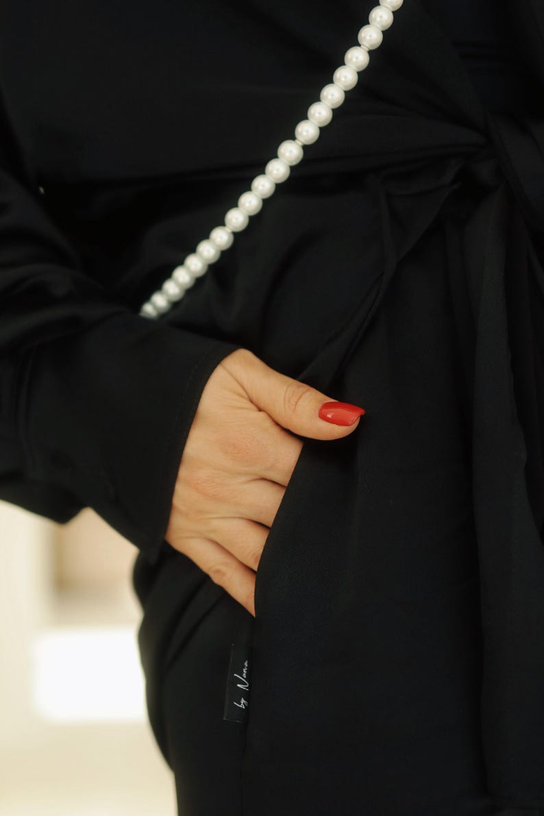May include: A black long-sleeved shirt with a pearl necklace and a pocket on the side. The shirt has a label that says "by Uma".