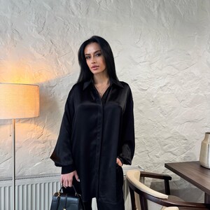 May include: A woman wearing a black satin shirt and matching trousers. She is holding a black leather handbag with a gold clasp.