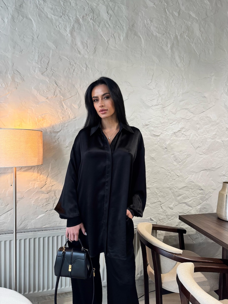 May include: A woman wearing a black satin shirt and matching trousers. She is holding a black leather handbag with a gold clasp.