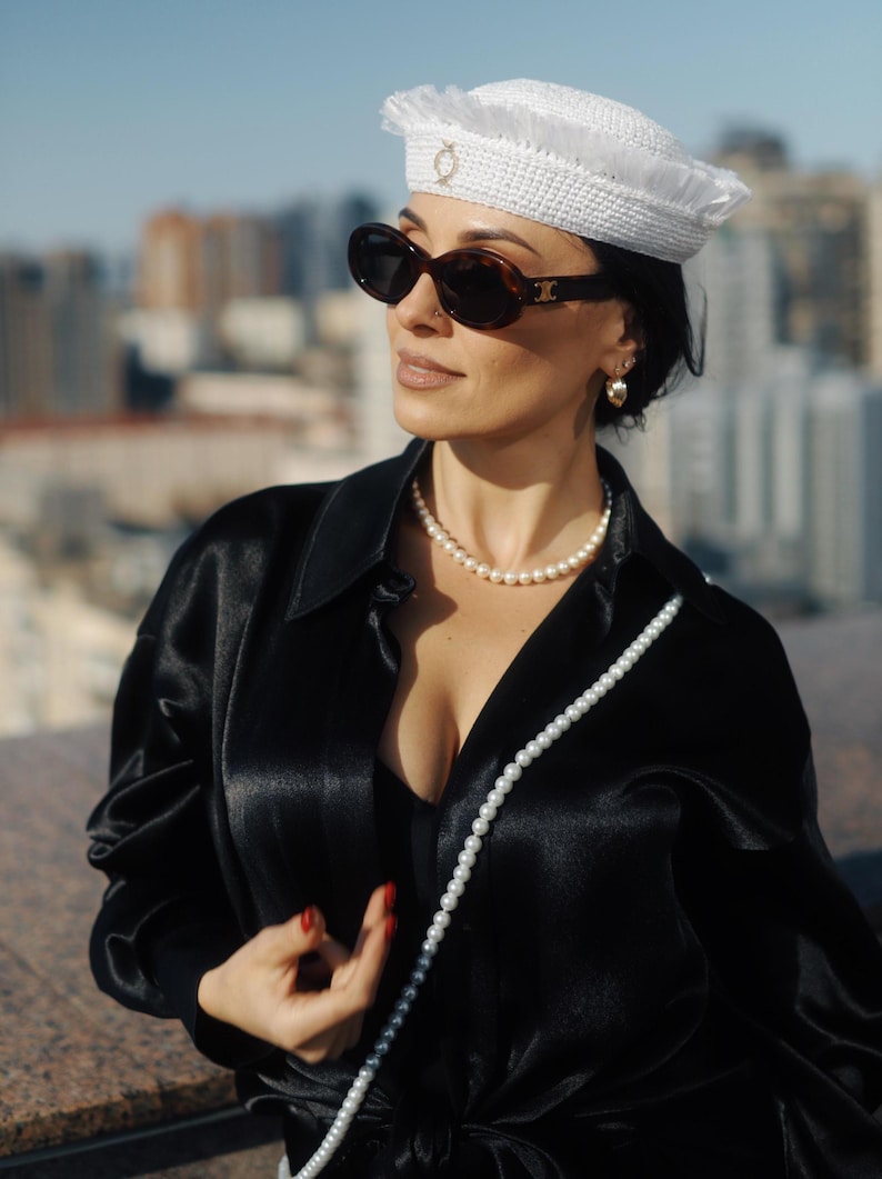 May include: A woman wearing a white crocheted hat with a gold accent, black sunglasses, a black satin shirt, and a long pearl necklace.