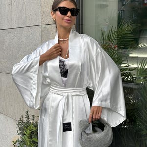 May include: A white satin robe with wide sleeves and a matching belt. The robe is worn over a white top with black lace detailing. A small gray handbag is held in the hand. The person is wearing sunglasses and a pearl necklace.