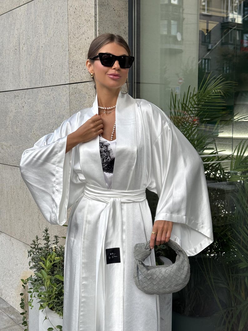 May include: A white satin robe with wide sleeves and a matching belt. The robe is worn over a white top with black lace detailing. A small gray handbag is held in the hand. The person is wearing sunglasses and a pearl necklace.