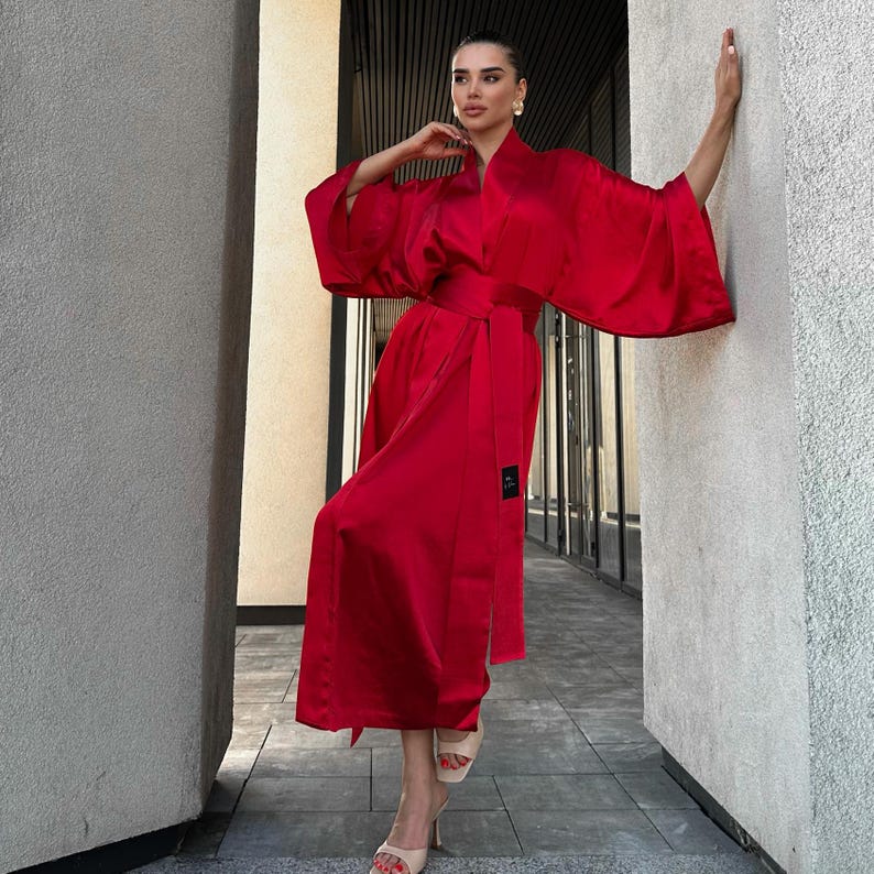 May include: A vibrant red satin robe with wide sleeves and a matching belt. The robe is knee-length and features a small label on the side. The model is wearing beige heels. The background is a neutral-toned outdoor setting.