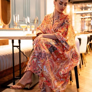 May include: A long, flowing dress with a vibrant paisley pattern in shades of orange, red, and black. The dress features a wrap-style design and is worn with brown heeled sandals. The setting appears to be a restaurant.