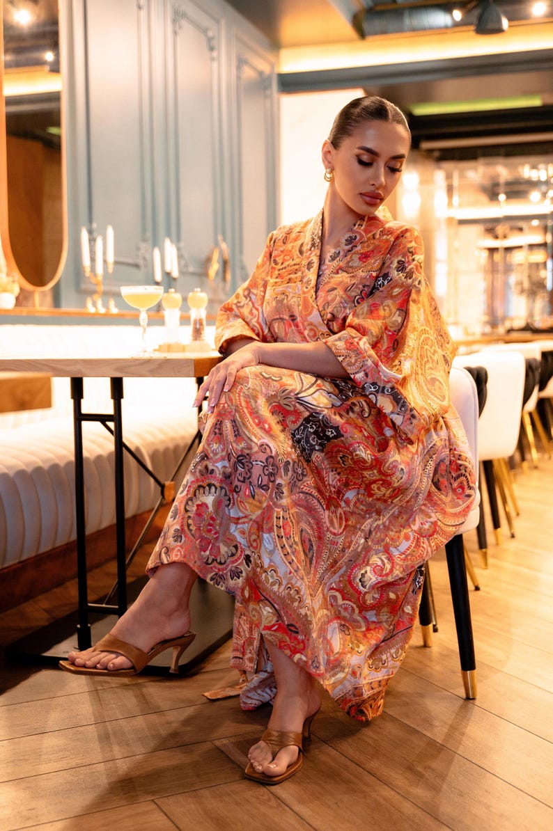 May include: A long, flowing dress with a vibrant paisley pattern in shades of orange, red, and black. The dress features a wrap-style design and is worn with brown heeled sandals. The setting appears to be a restaurant.