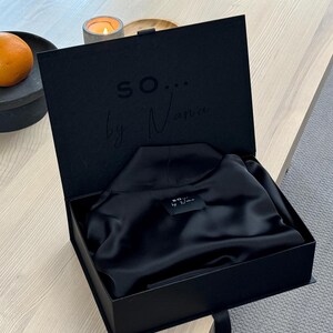 May include: A black presentation box, open to reveal a folded black garment. The box lid has the text "so... by Nana" embossed inside. An orange sits on a black plate in the background.