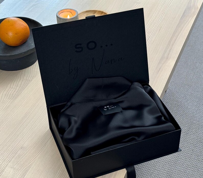 May include: A black presentation box, open to reveal a folded black garment. The box lid has the text "so... by Nana" embossed inside. An orange sits on a black plate in the background.