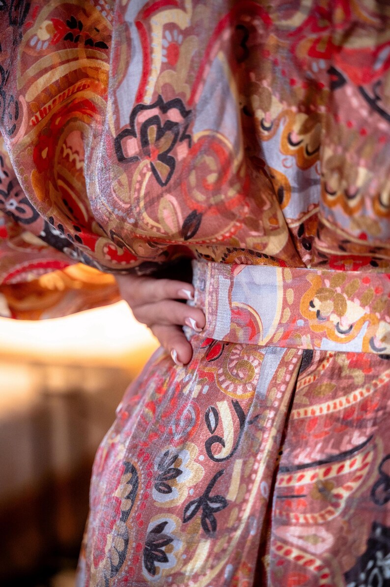 May include: A patterned garment in red, brown, and gold tones, featuring a paisley design with floral and abstract shapes. The garment, possibly a robe or kimono, has a belt. The fabric is visible in close-up.