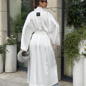 May include: A white, long-sleeved robe with a matching belt. The robe has a square black label near the neckline. The person is wearing blue jeans and black shoes. A gray handbag is held in the left hand.