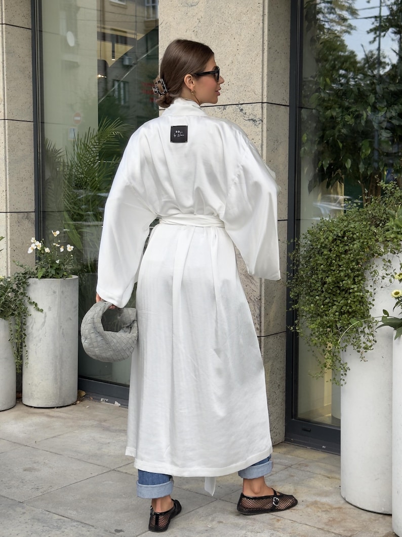 May include: A white, long-sleeved robe with a matching belt. The robe has a square black label near the neckline. The person is wearing blue jeans and black shoes. A gray handbag is held in the left hand.