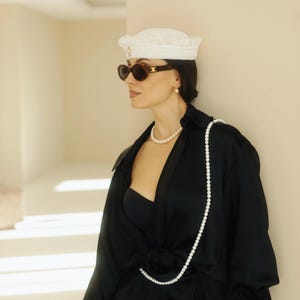 May include: A woman wearing a white crocheted hat with a gold accent, black sunglasses, a black satin shirt, and a long pearl necklace. The shirt is open at the front, revealing a black bandeau top. The woman is standing in a hallway with white walls and a white archway.