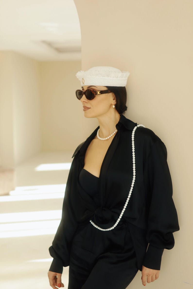 May include: A woman wearing a white crocheted hat with a gold accent, black sunglasses, a black satin shirt, and a long pearl necklace. The shirt is open at the front, revealing a black bandeau top. The woman is standing in a hallway with white walls and a white archway.