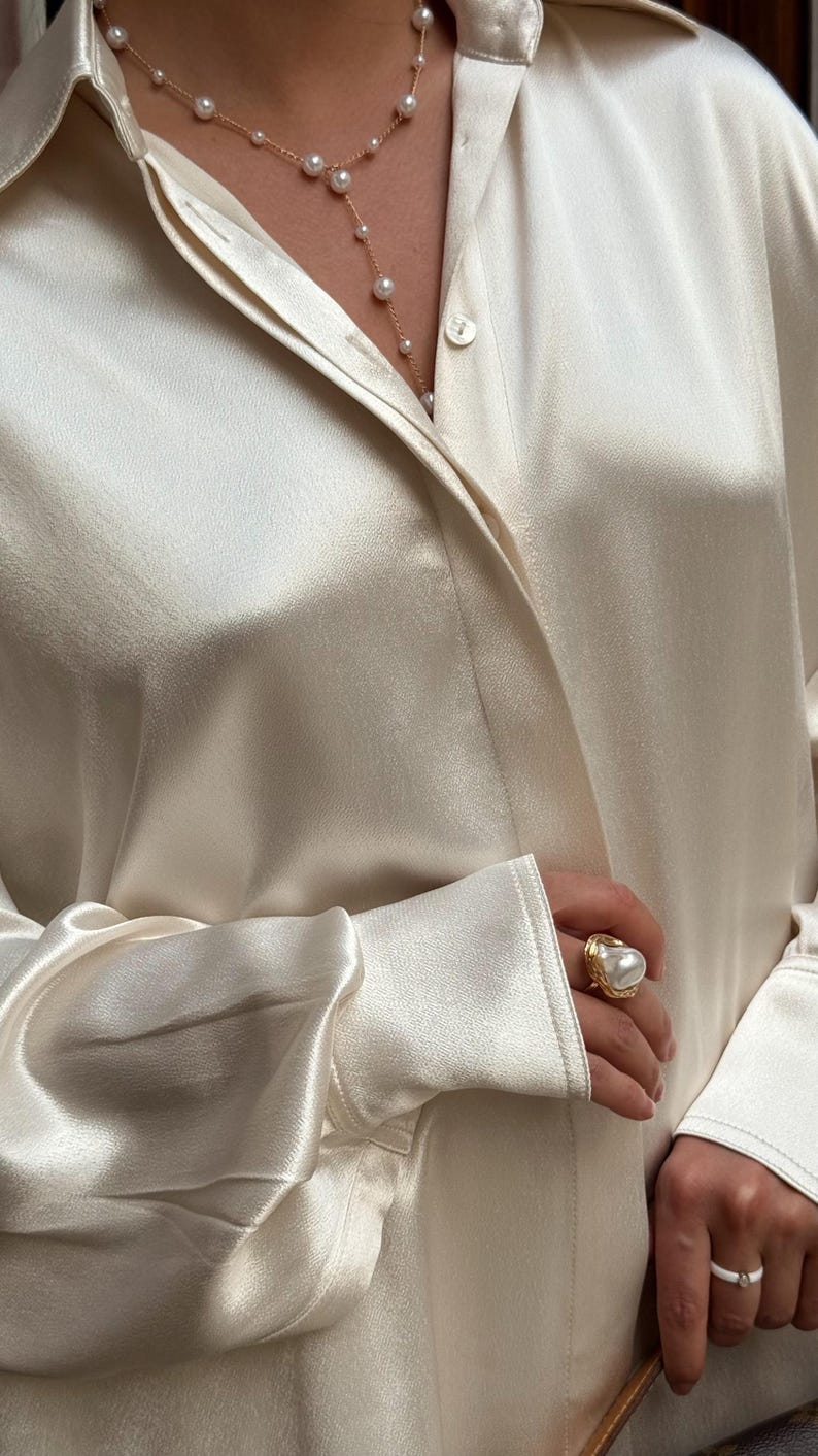 May include: Cream-colored silk blouse with a gold pearl necklace and a large pearl ring. The ring has a gold band with a textured design. The blouse has long sleeves and a button-down front.