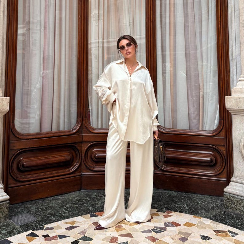 May include: Cream-colored satin two-piece set, featuring a long-sleeved button-down shirt and wide-leg pants. The outfit is worn with sunglasses and a necklace. A brown handbag is held in the hand. The background includes a dark wooden door and a patterned floor.