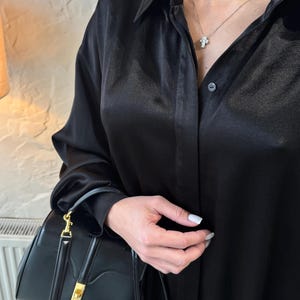 May include: A black satin shirt with a button-up front and long sleeves. The shirt is being worn by a person holding a black leather handbag with a gold clasp.