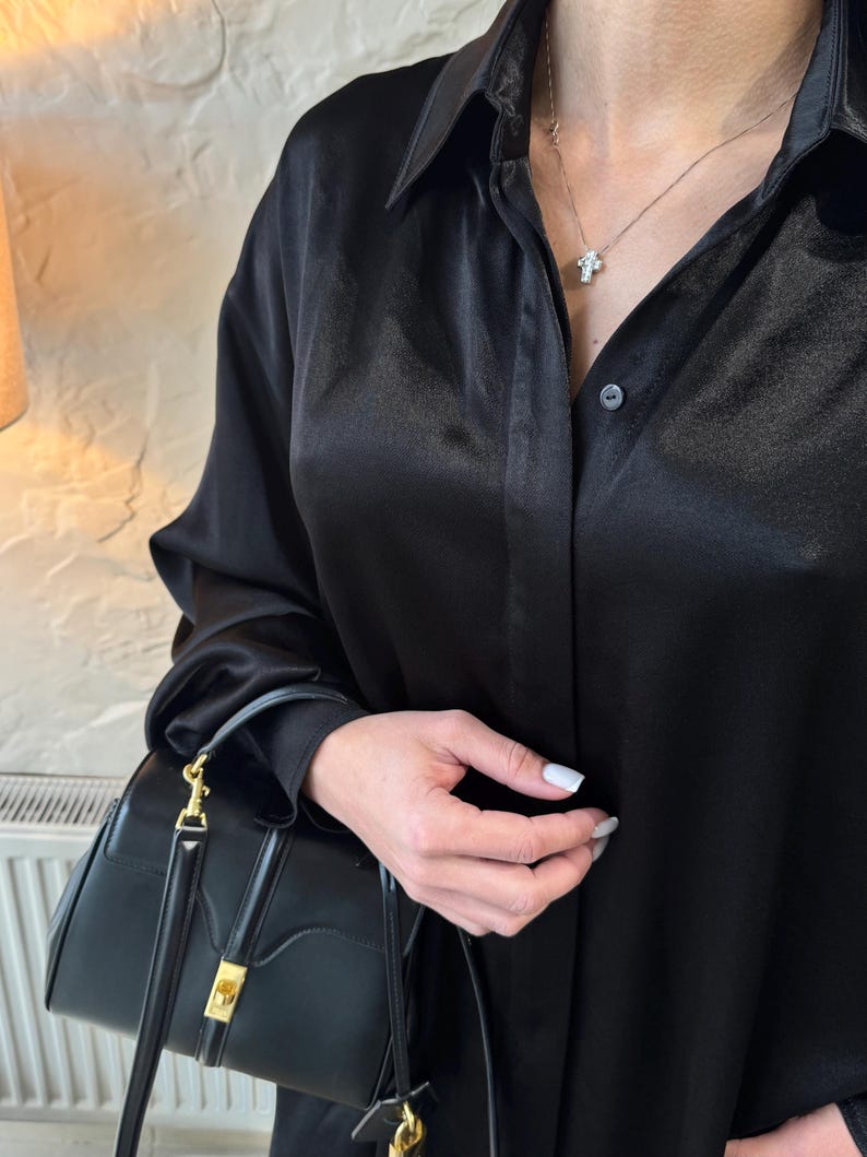 May include: A black satin shirt with a button-up front and long sleeves. The shirt is being worn by a person holding a black leather handbag with a gold clasp.